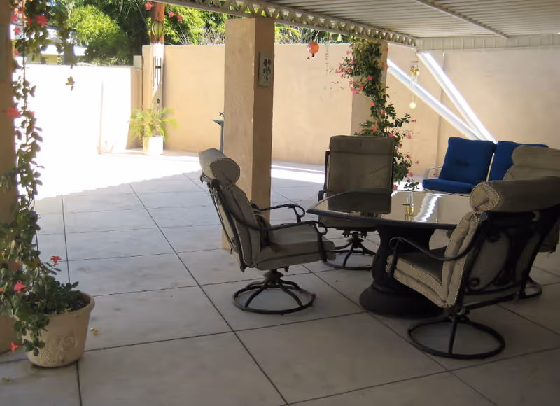 Covered outdoor patio area with a round glass-top table surrounded by four cushioned swivel chairs. There are potted plants with flowers hanging from the ceiling and climbing a pillar. In the background, there is a blue cushioned bench against a beige wall.