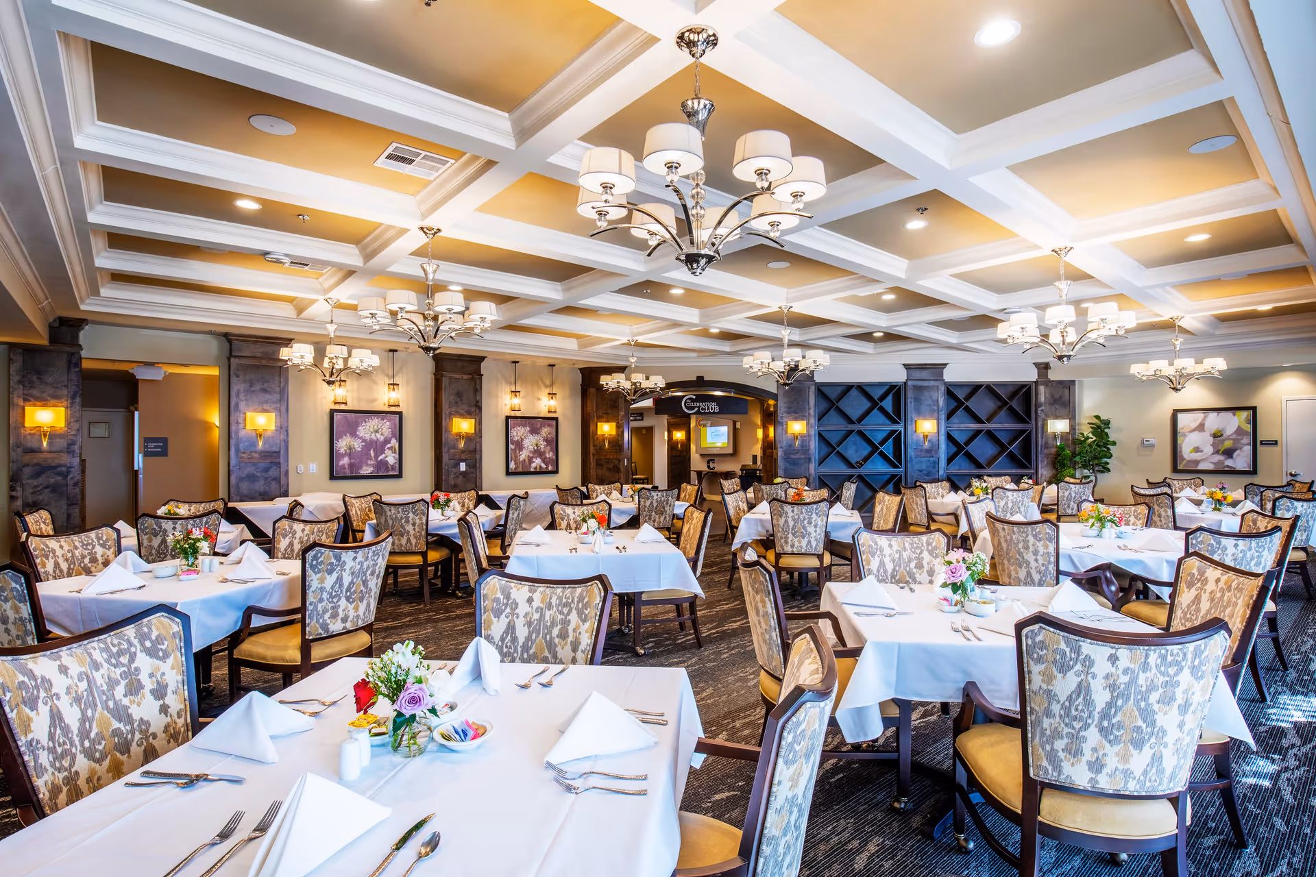 A spacious and elegant dining room with multiple tables covered in white tablecloths, each set with folded napkins, silverware, and small floral centerpieces. The room features patterned upholstered chairs, warm lighting from chandeliers and wall sconces, decorative ceiling beams, and framed floral artwork on the walls.