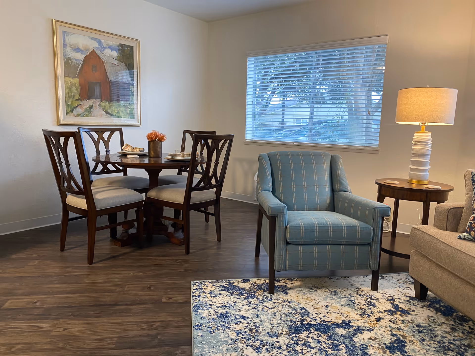 A cozy living and dining area featuring a round wooden dining table with four cushioned chairs, a blue patterned armchair, a wooden side table with a lamp, and a large window with blinds. A framed painting of a red barn hangs on the wall above the dining table, and a blue and white patterned rug is partially visible on the floor.