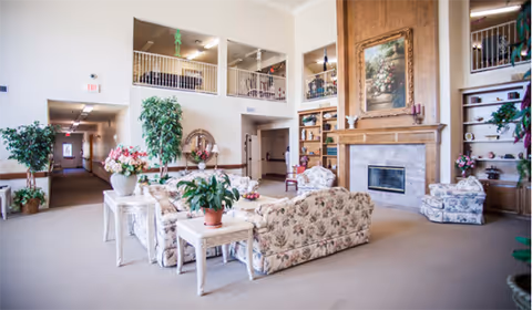 Bright senior living lobby with floral sofas, a fireplace, plants, and a second-floor balcony.