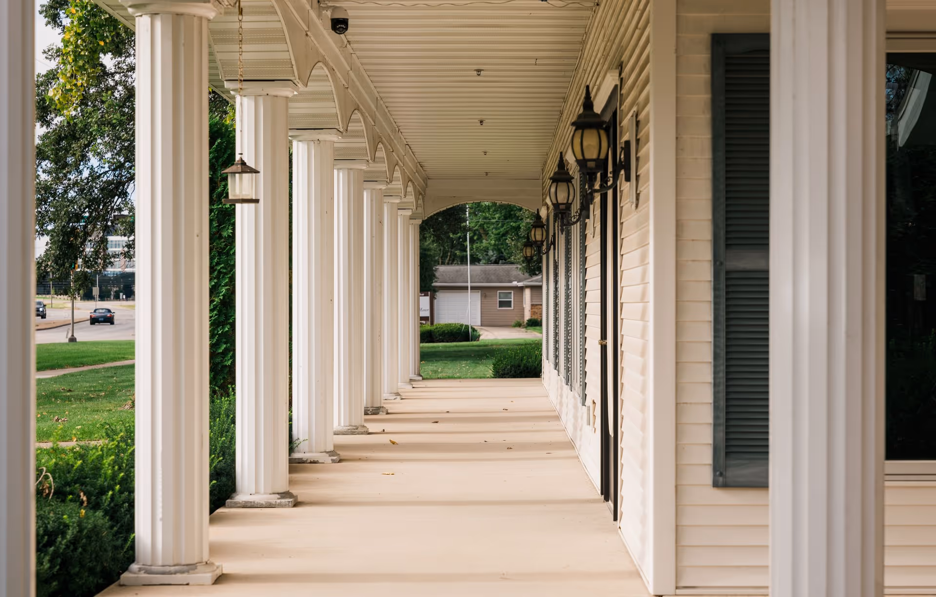 A covered outdoor walkway with white columns and beige siding on the right side, featuring black lantern-style wall lights and green bushes along the left side. Trees and a road with cars are visible in the background.