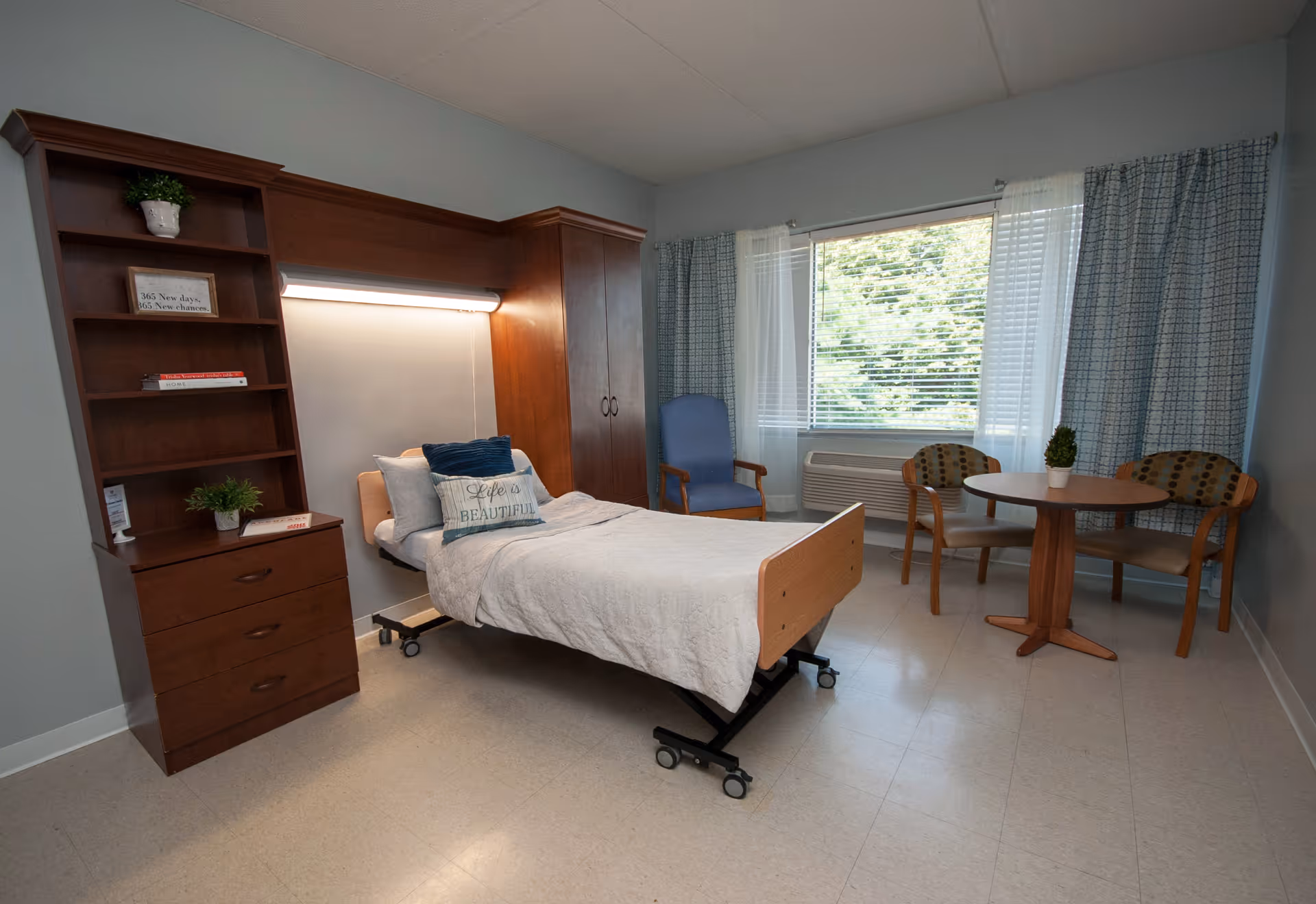 A senior living facility bedroom with a single adjustable bed featuring pillows and a blanket. To the left of the bed is a wooden shelving unit with drawers and decorative plants. A large window with curtains lets in natural light. Near the window, there is a small round table with two chairs and a small plant on the table. The room has light-colored walls and flooring.