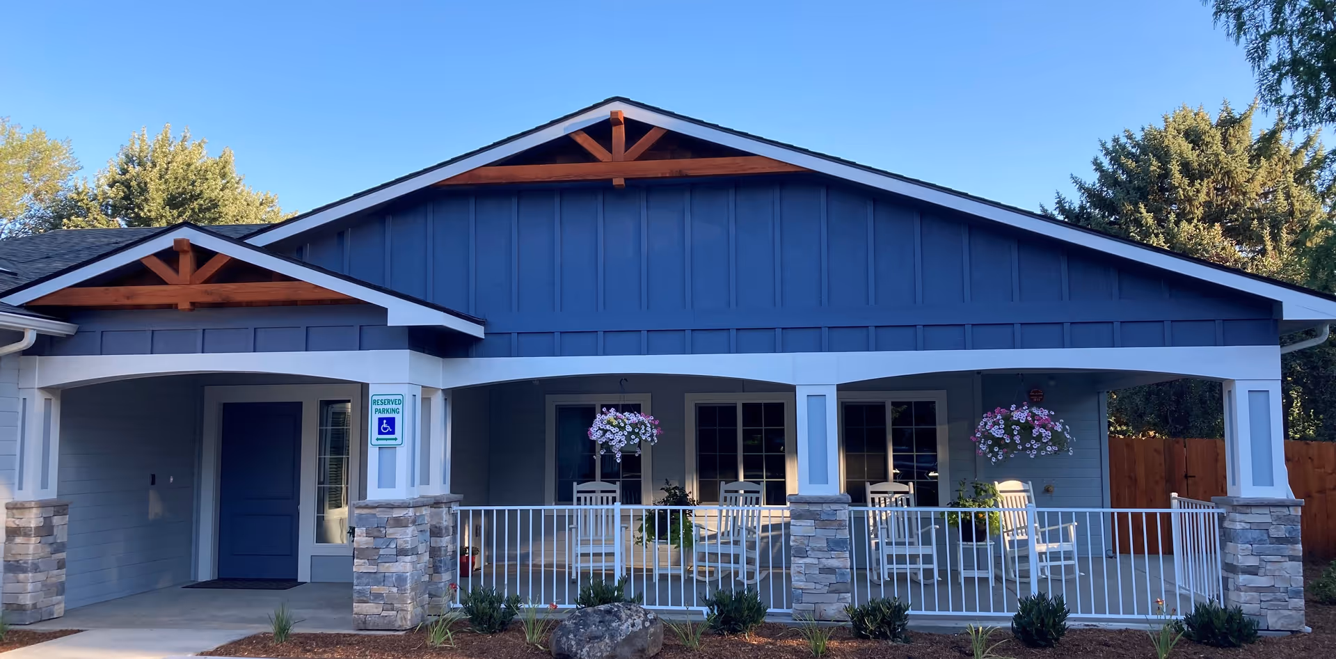 Front exterior view of a single-story building with blue siding and white trim. The building has a covered porch with white railings, stone pillars, hanging flower baskets, and several white rocking chairs. There is a reserved parking sign for handicapped parking near the entrance. Trees and a wooden fence are visible in the background under a clear blue sky.