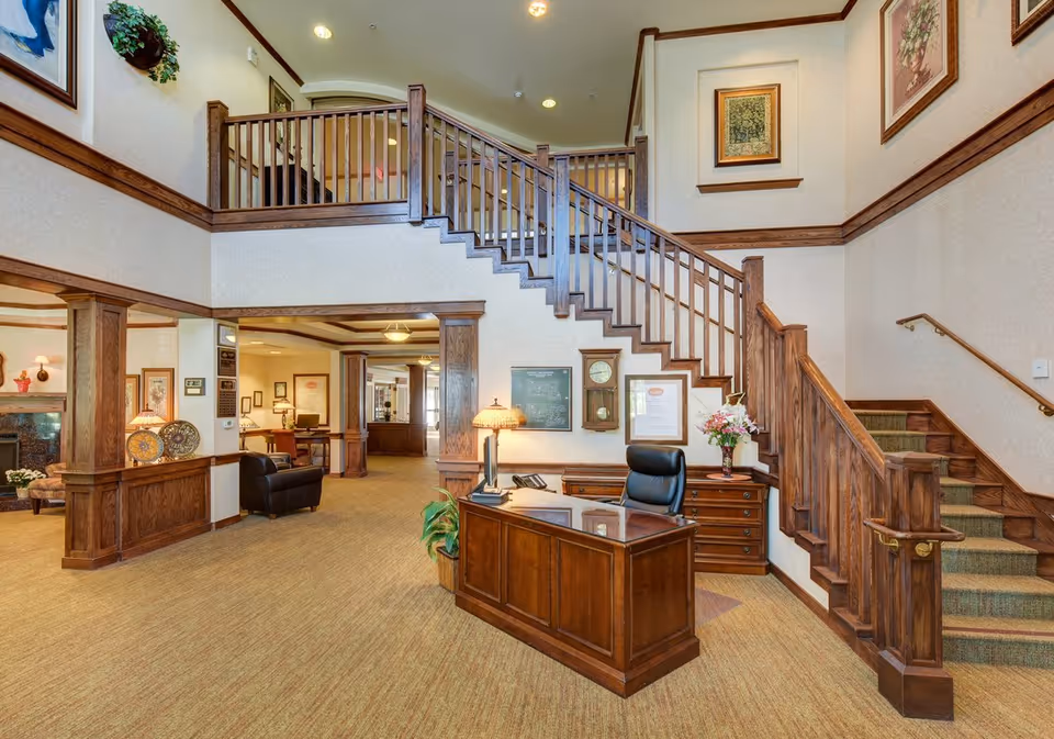 Interior view of a senior living facility lobby with a wooden reception desk and chair in front of a staircase with wooden railings. The walls are decorated with framed artwork and a clock. There is a lamp and a vase with flowers on the desk. The area is carpeted and well-lit with ceiling lights.