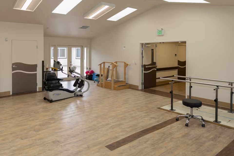 A spacious rehabilitation room with light wood flooring and white walls, featuring exercise equipment including a recumbent bike, parallel bars for walking practice, a small set of stairs with handrails, and a black rolling stool. The room has large windows and doors letting in natural light, and an exit sign above a double door leading to a hallway.