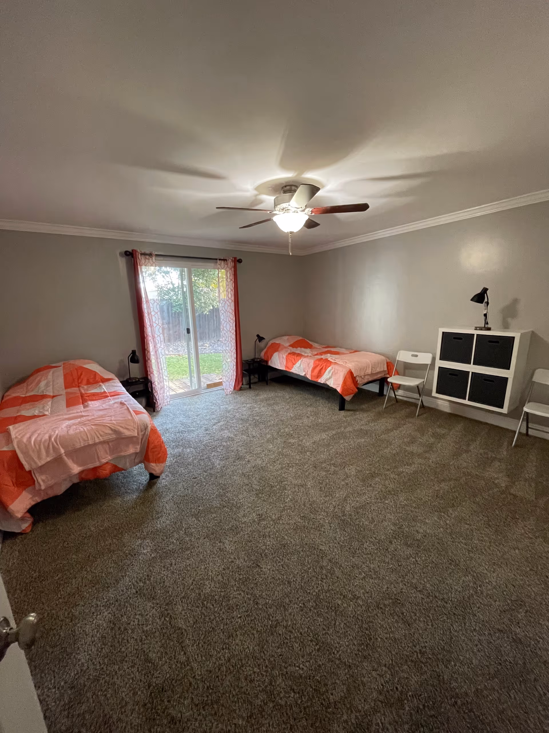 A bedroom with two single beds covered in orange and pink bedding. The room has gray carpet, light gray walls, a ceiling fan with light, and a sliding glass door with red patterned curtains leading outside. There are two small black nightstands with lamps next to each bed, two white folding chairs, and a white wall-mounted storage unit with black bins.