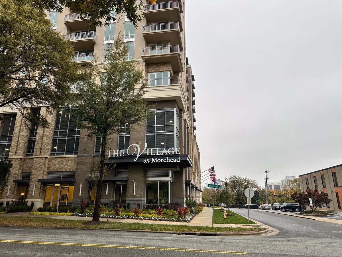 Front exterior of a multi-story building labeled 'The Village on Morehead' with trees, a sidewalk, and an American flag.