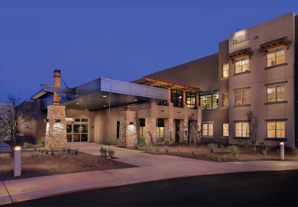 Exterior view of a modern senior living facility building at dusk with stone pillars supporting a covered entrance, illuminated windows, and landscaped surroundings.