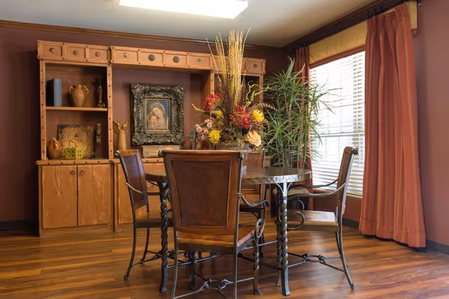 A cozy interior room with a round glass-top table surrounded by four wooden chairs with woven backs. On the table is a large floral arrangement with various flowers and tall grasses. Behind the table is a wooden cabinet with shelves displaying pottery and a framed picture. To the right, there is a window with blinds and long rust-colored curtains. The floor is wooden, and the walls are painted a warm brown color.