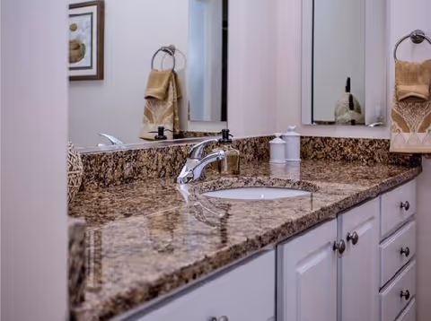 A bathroom countertop with a granite surface, featuring a built-in sink with a chrome faucet. On the countertop are soap dispensers and a decorative item. Two beige hand towels hang on towel rings on either side of the mirror above the sink. White cabinetry with silver knobs is below the countertop.