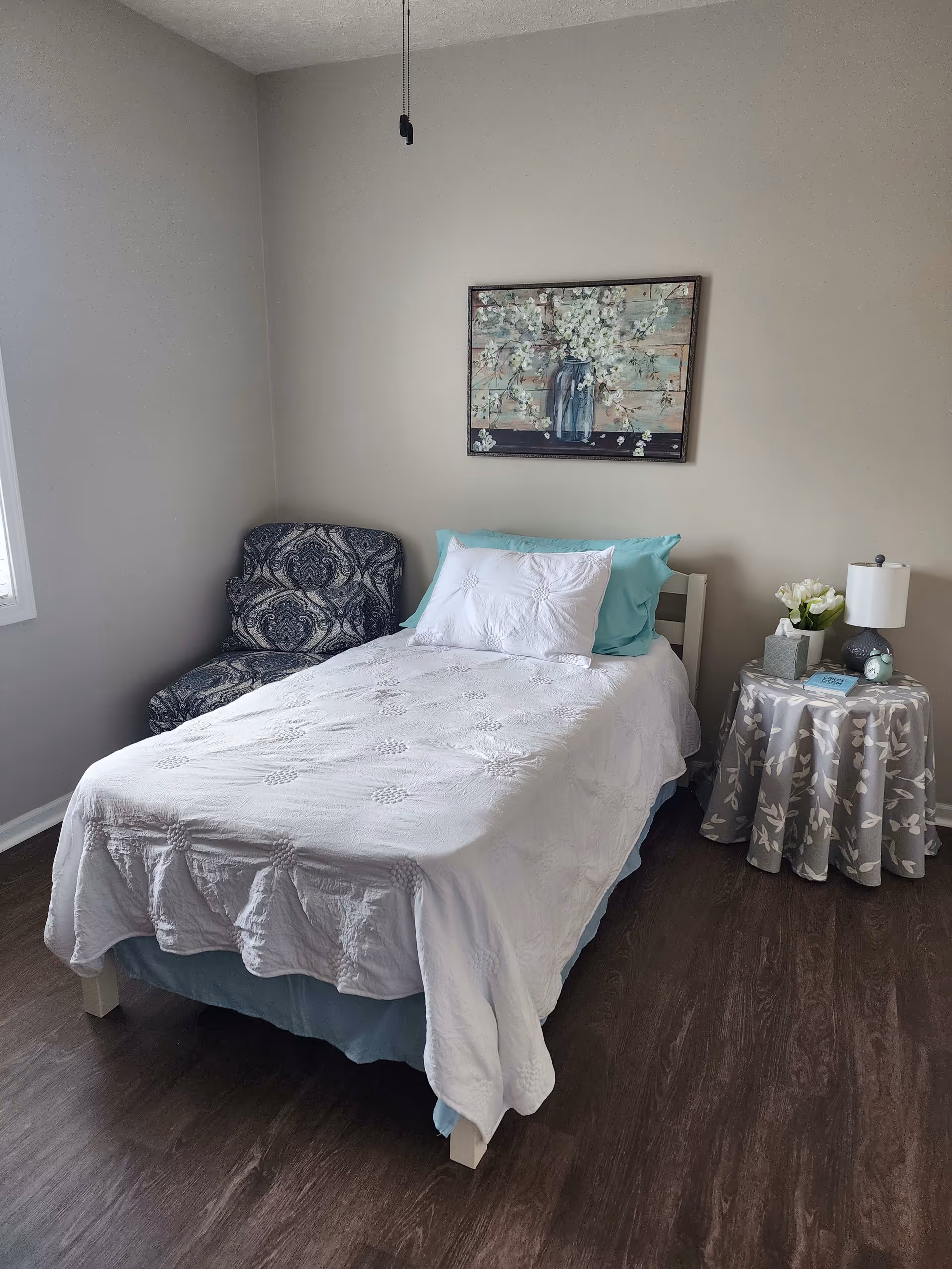 A small bedroom with a single bed covered in a white quilt and light blue pillows. To the left of the bed is a patterned armchair, and to the right is a round side table with a floral tablecloth, a lamp, a small clock, a tissue box, and a vase with white flowers. A painting of white flowers in a vase hangs on the beige wall above the bed. The floor is dark wood, and there is a window on the left wall.
