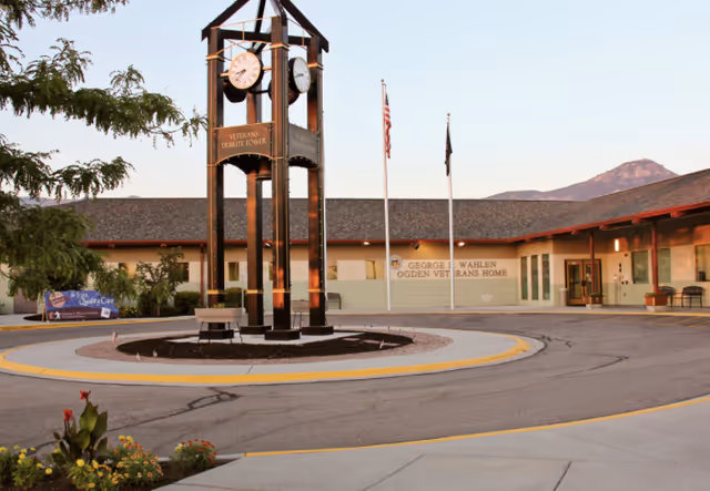 Exterior view of George E. Wahlen Ogden Veterans Home featuring a circular driveway with a Veterans Tribute Tower clock structure in the center, two flagpoles with American and POW/MIA flags, and a single-story building with a mountain in the background.