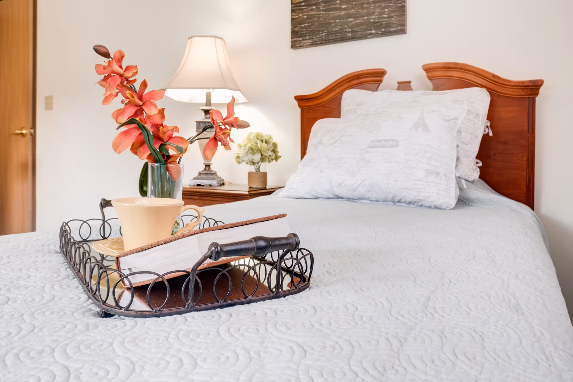 A neatly made bed with a wooden headboard, white patterned pillows, and a light blue quilted bedspread. On the bed is a decorative tray holding a book, a beige cup and saucer, and a vase with pink flowers. In the background, there is a nightstand with a lamp and a small plant, and a closed wooden door.