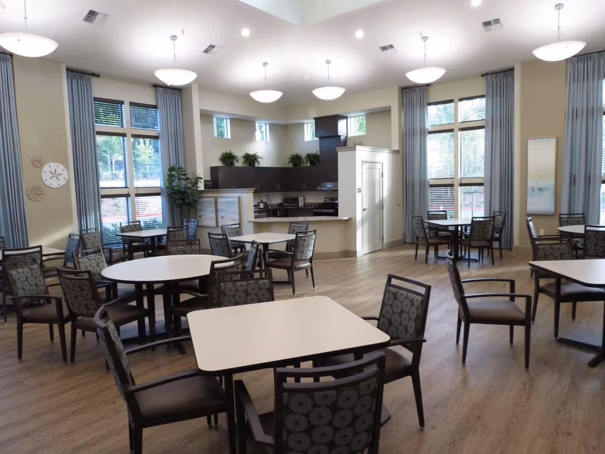 A spacious dining room with multiple round and square tables surrounded by patterned chairs. The room has large windows with blue curtains allowing natural light to fill the space. In the background, there is a kitchen area with dark cabinets, a stove, and a countertop. The floor is wooden, and the ceiling has several round light fixtures.