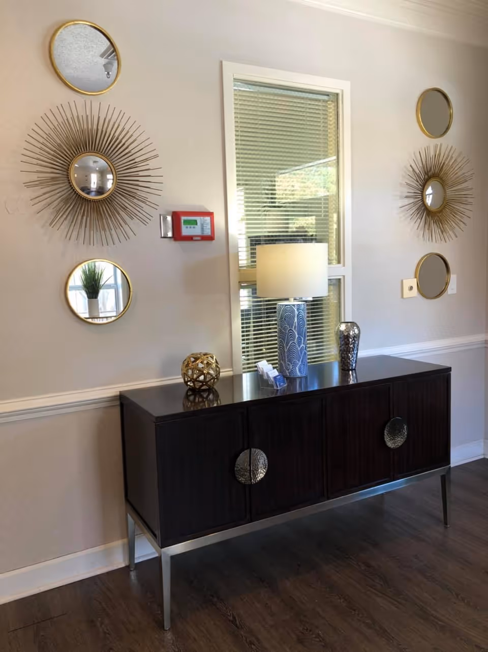 A dark wooden console table with metallic circular handles stands against a light-colored wall. On top of the table are a blue and white patterned lamp with a white shade, a decorative metallic vase, a gold geometric decorative object, and some business cards. The wall behind the table features a window with blinds and six decorative round mirrors with gold frames, two of which have sunburst designs. A red fire alarm is mounted on the wall to the left of the window.