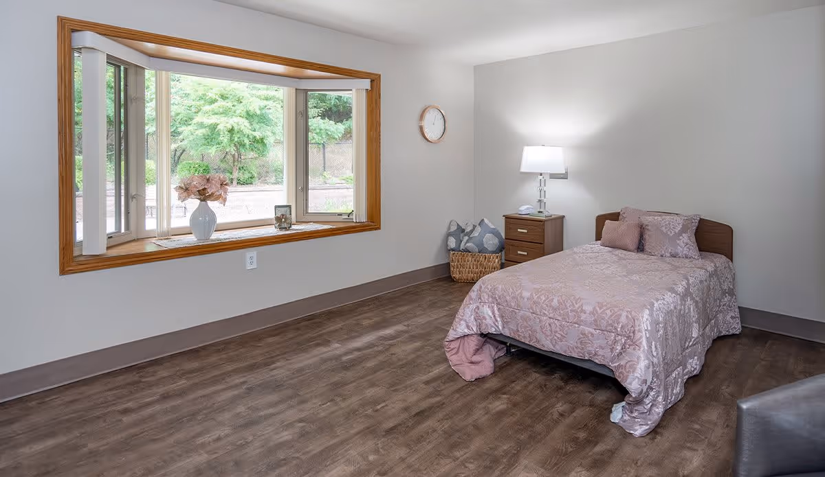 A simple, clean bedroom with a single bed covered in a pink patterned bedspread and matching pillows. Next to the bed is a wooden nightstand with a lamp and a telephone. A basket with pillows is placed in the corner. A large window with a wooden frame shows a view of green trees outside. The room has wood flooring and light-colored walls.