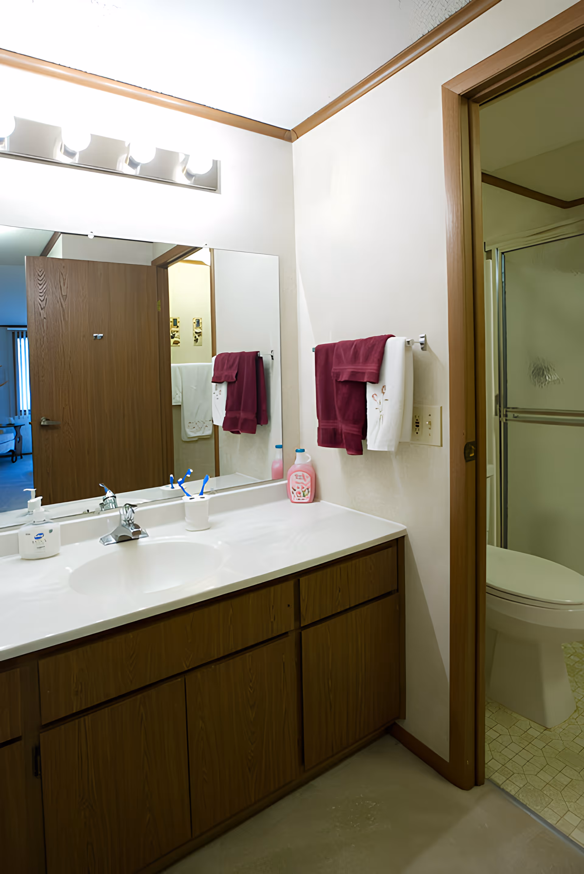 A bathroom vanity with a white countertop, a sink, and wooden cabinets below. On the countertop are a soap dispenser, a cup with toothbrushes, and a bottle of pink liquid soap. Above the vanity is a large mirror with four bright lights mounted above it. To the right, there is a towel rack with a burgundy towel and a white towel hanging. Through an open doorway, a toilet and a shower with sliding glass doors are visible. The bathroom has light-colored walls and a beige tiled floor in the shower area.