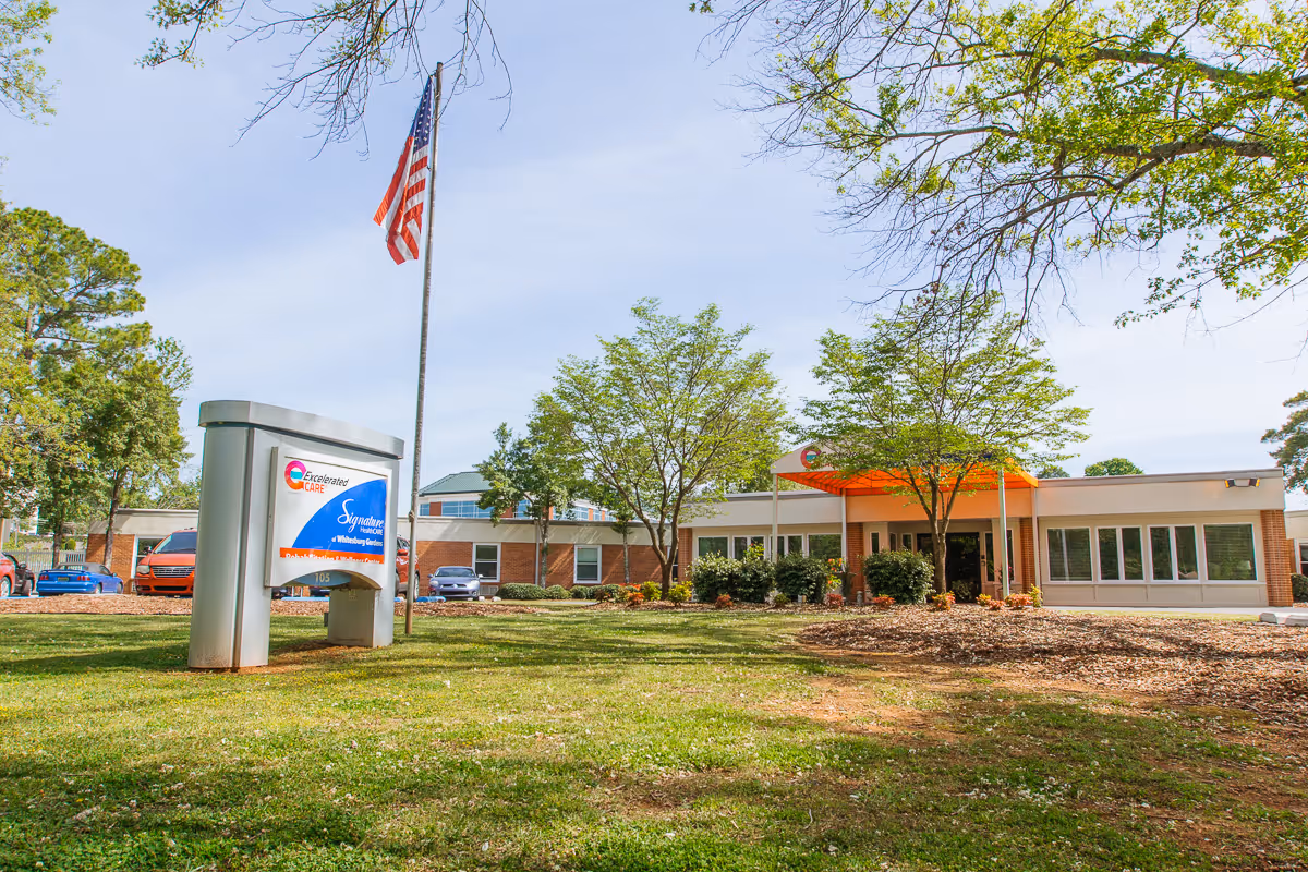 Exterior view of Rocket City Rehabilitation and Healthcare Center building with a lawn, trees, an American flag on a flagpole, and a sign in front displaying the facility's name and logo.