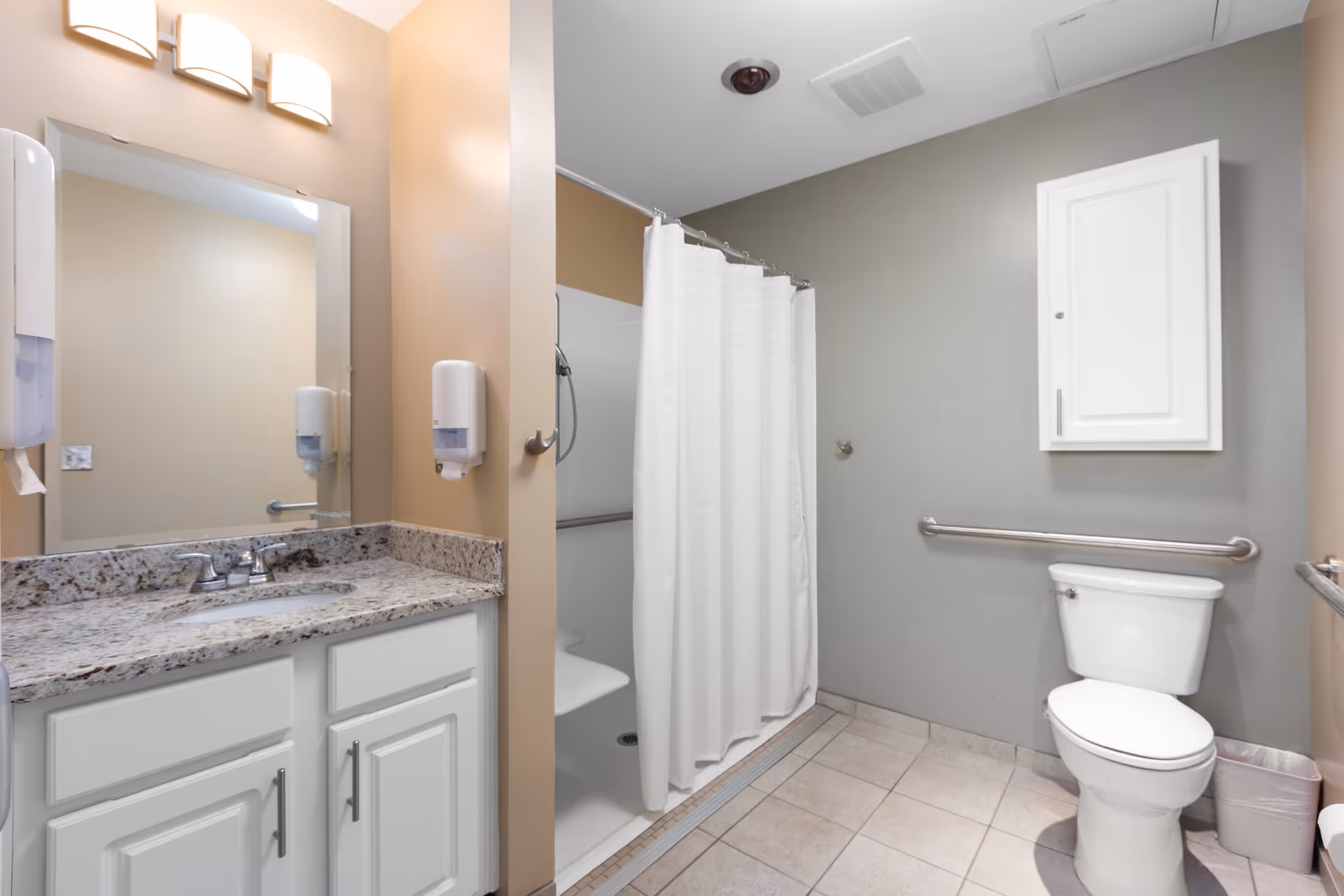 A clean and accessible bathroom with a white toilet, a shower area with a white curtain and a built-in seat, a granite countertop with a sink, a large mirror, and beige and gray walls. There are grab bars near the toilet and inside the shower for safety.