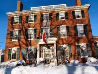 Three-story red brick building with green shutters, a central entrance with a flag, and snow-covered landscaping with tree shadows on the facade.