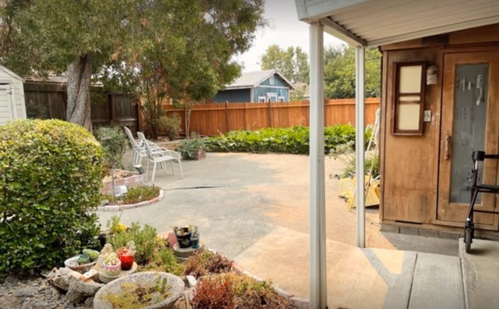 A backyard patio with potted plants and garden beds, white metal chairs, a wooden side door under a covered porch, and a fenced yard with trees.