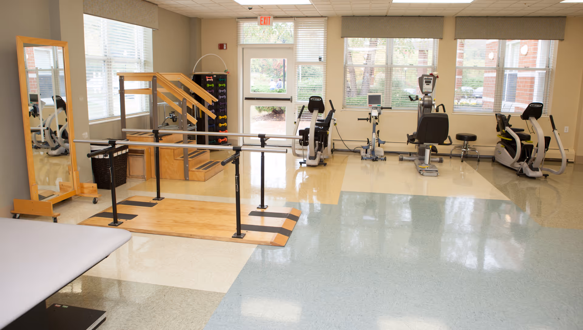 A bright physical therapy or exercise room with large windows letting in natural light. The room contains parallel bars for walking exercises, a set of wooden stairs with handrails, a rack of colorful dumbbells, and several exercise machines including stationary bikes and a rowing machine. There is a full-length mirror on the left side and a treatment table partially visible in the foreground.