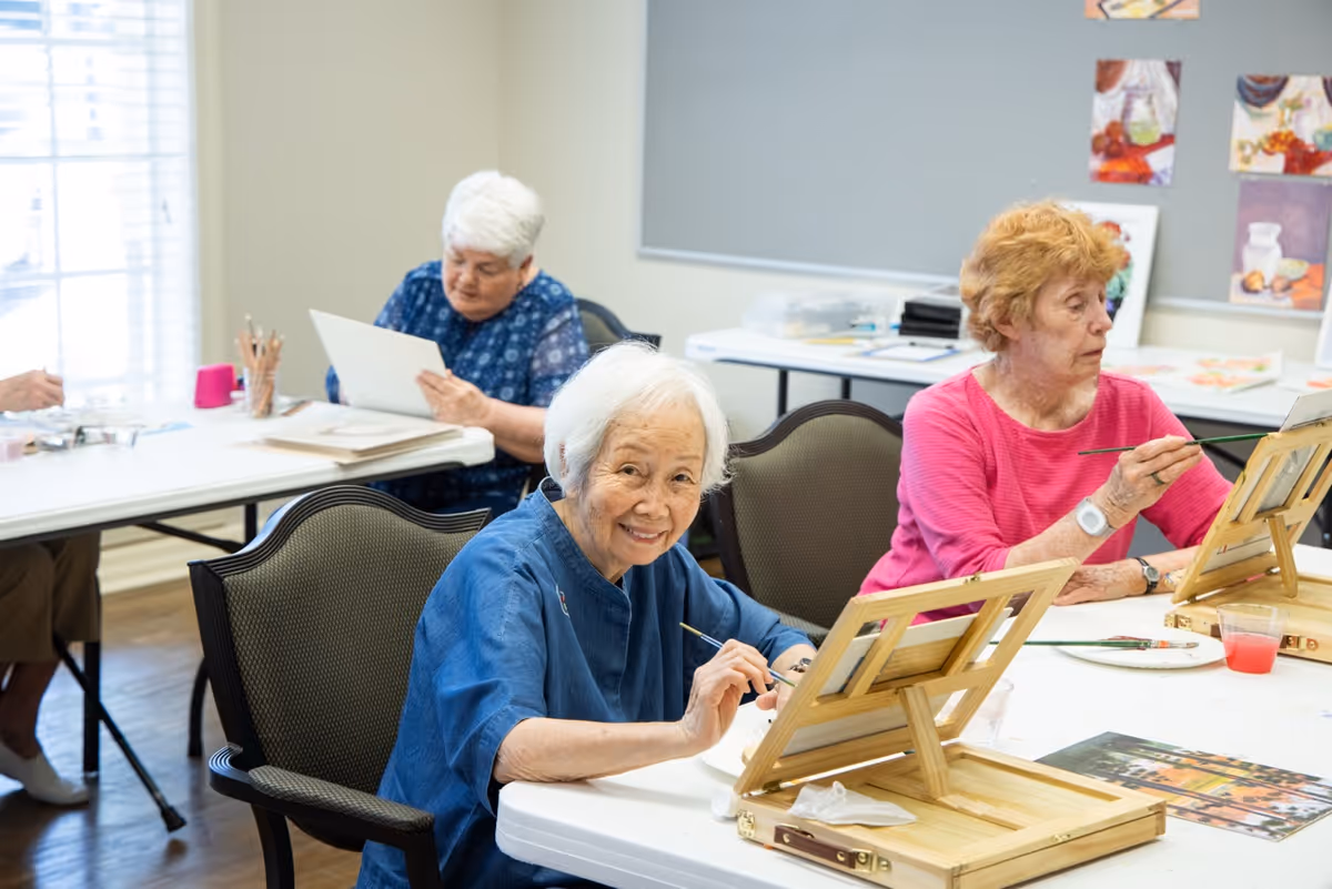 Three elderly women seated at tables in a bright room, engaged in painting and drawing activities. One woman in the foreground smiles at the camera while holding a paintbrush and working on a canvas on a wooden easel. The other two women are focused on their artwork, with one painting and the other drawing on paper. The room has light-colored walls and artwork displayed on a bulletin board in the background.