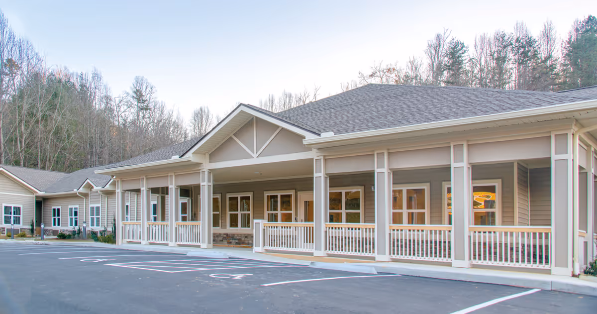Exterior view of a single-story senior living facility building with a covered porch, multiple windows, and a parking lot with handicap spaces in front. Trees and a clear sky are visible in the background.