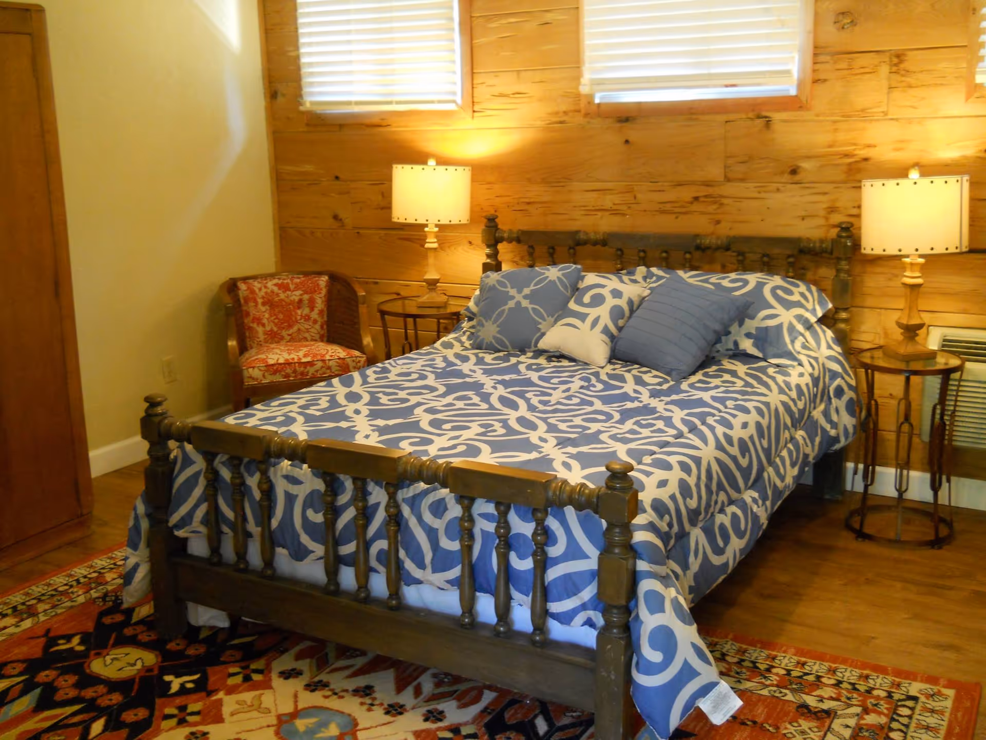A cozy bedroom with a wooden bed frame and a blue and white patterned bedspread. There are three decorative pillows on the bed. The room has wooden walls with two windows covered by blinds. On either side of the bed are round wooden nightstands with matching lamps. A red patterned armchair is placed in the corner next to a wooden wardrobe. The floor is wooden with a colorful patterned rug partially visible under the bed.