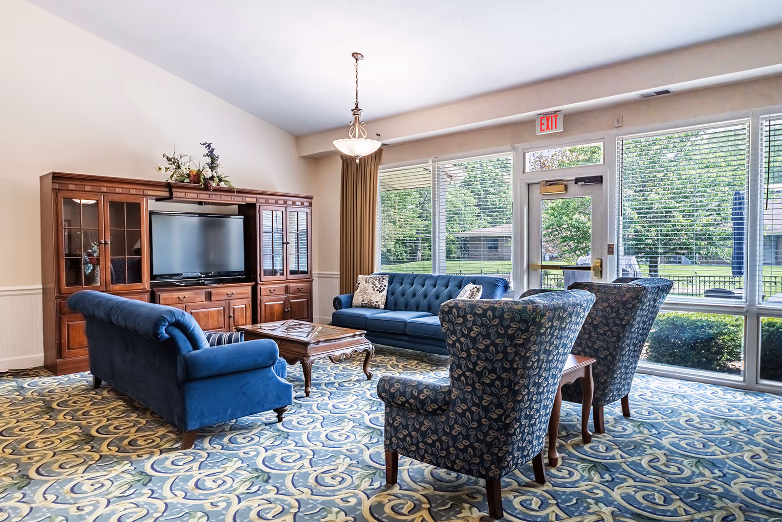A bright senior living facility common area with large windows letting in natural light. The room features a blue sofa, two patterned armchairs, a wooden coffee table, and a wooden entertainment center with a flat-screen TV. The carpet has a blue and beige swirling pattern, and there is an exit door with a red exit sign above it.