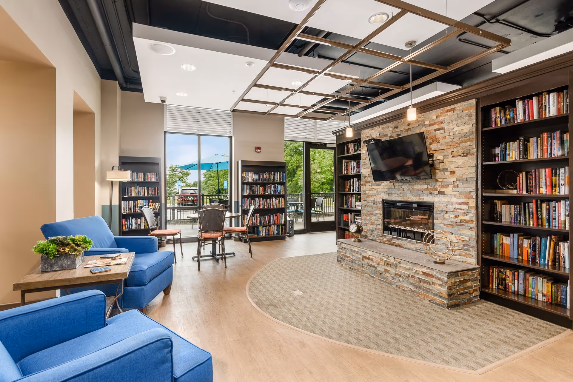 Bright communal lounge with blue armchairs, bookshelves, a stone fireplace with mounted TV, and seating by glass doors opening to an outdoor patio.