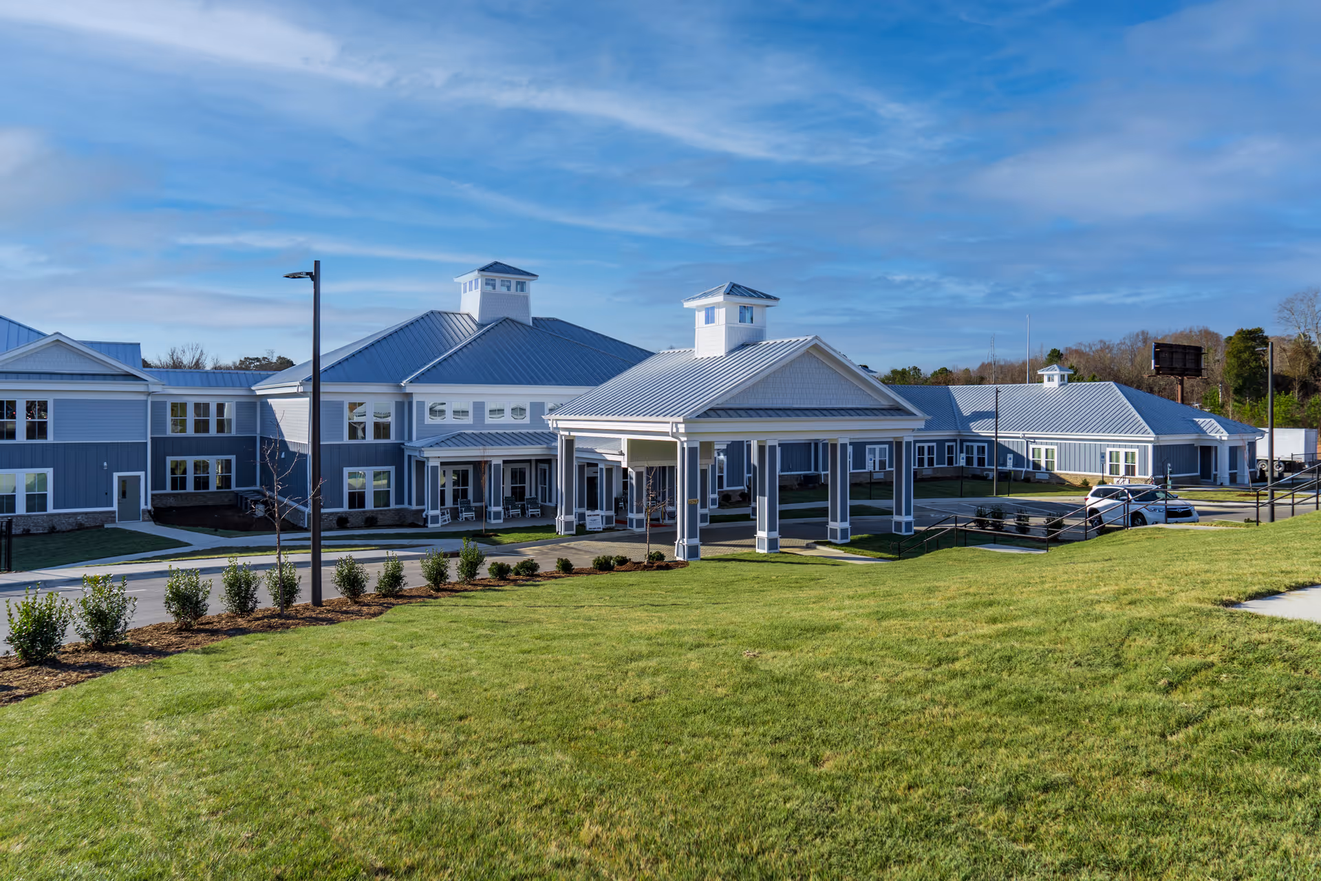 Exterior view of Arbor Ridge at Huntersville, a large senior living facility with gray siding and white trim. The building features multiple peaked roofs with cupolas, a covered entrance supported by columns, and a well-maintained lawn in the foreground under a partly cloudy blue sky.
