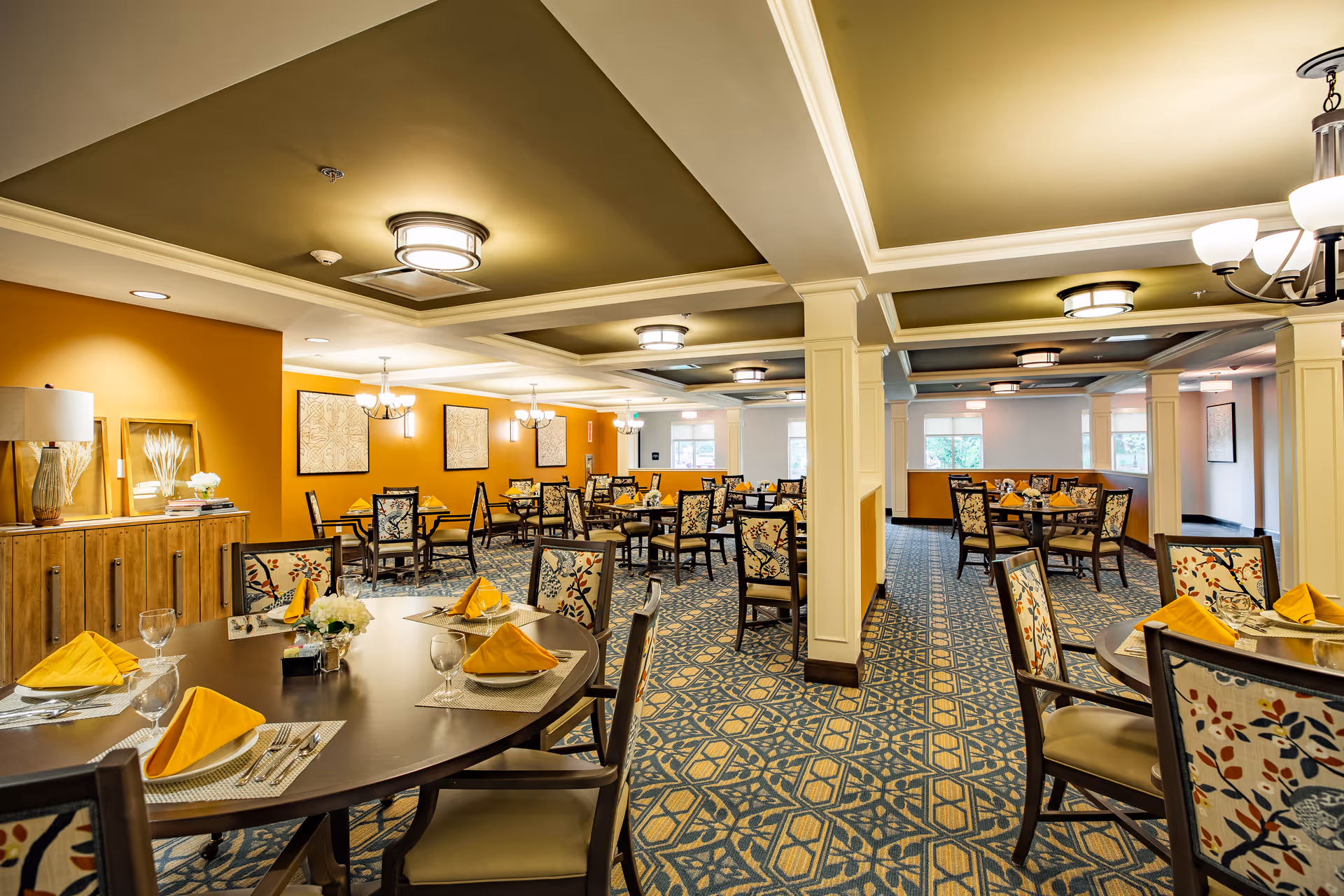 Bright, spacious dining room with round tables set with yellow napkins and floral-upholstered chairs.
