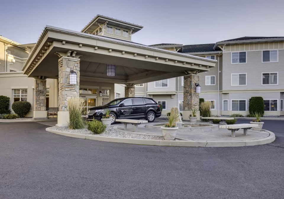 Entrance of a senior living facility with a covered drop-off area supported by stone pillars. A black vehicle with the Fairwinds logo is parked under the canopy. The building is a multi-story structure with beige siding and multiple windows. There are landscaped plants and benches around the circular driveway.