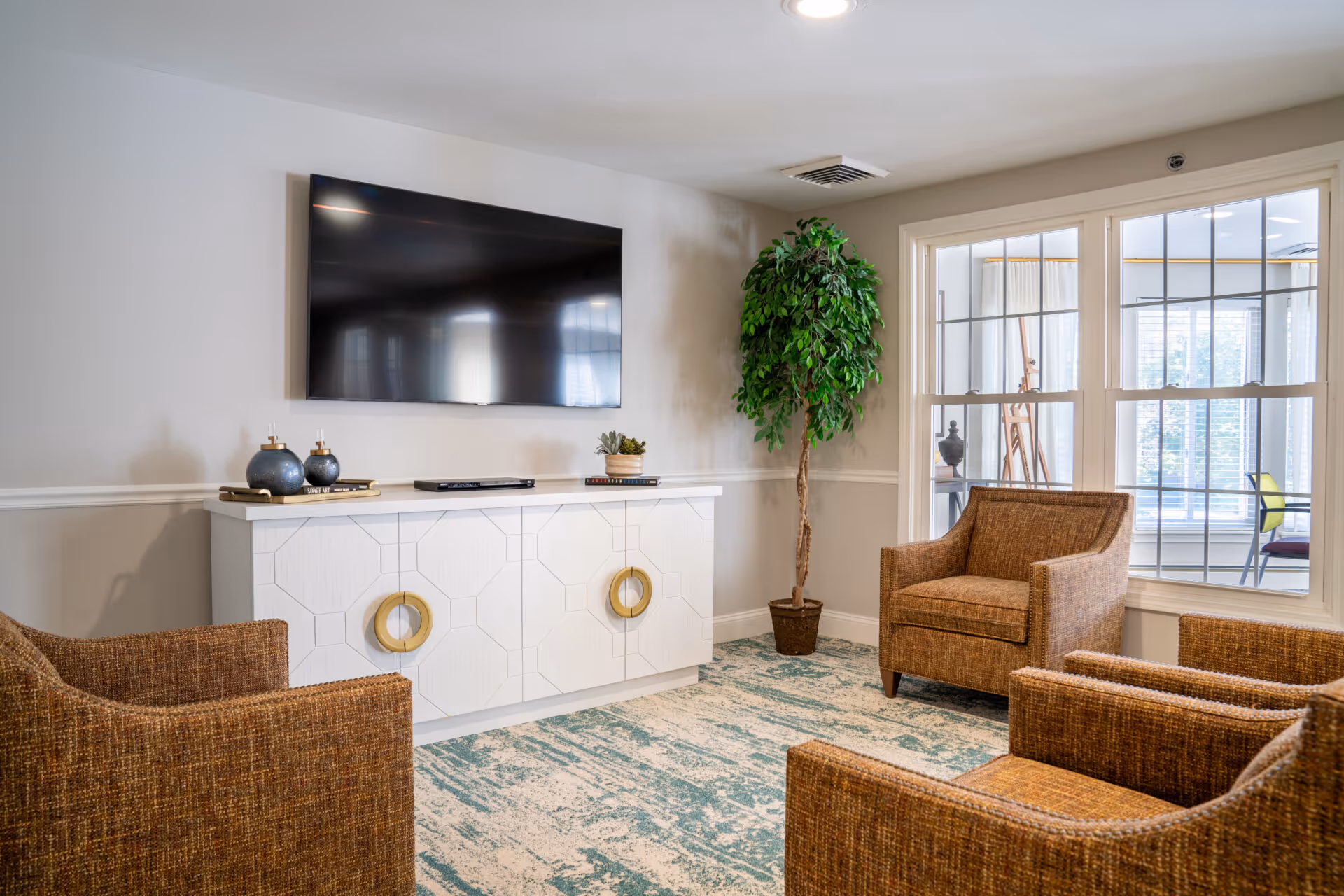 A cozy living room area with four brown woven armchairs arranged around a white cabinet with gold circular handles. A large flat-screen TV is mounted on the wall above the cabinet. A tall potted green plant is placed in the corner near a window with multiple panes, through which another room with chairs and an easel is visible. The room has light-colored walls and a patterned blue and beige carpet.