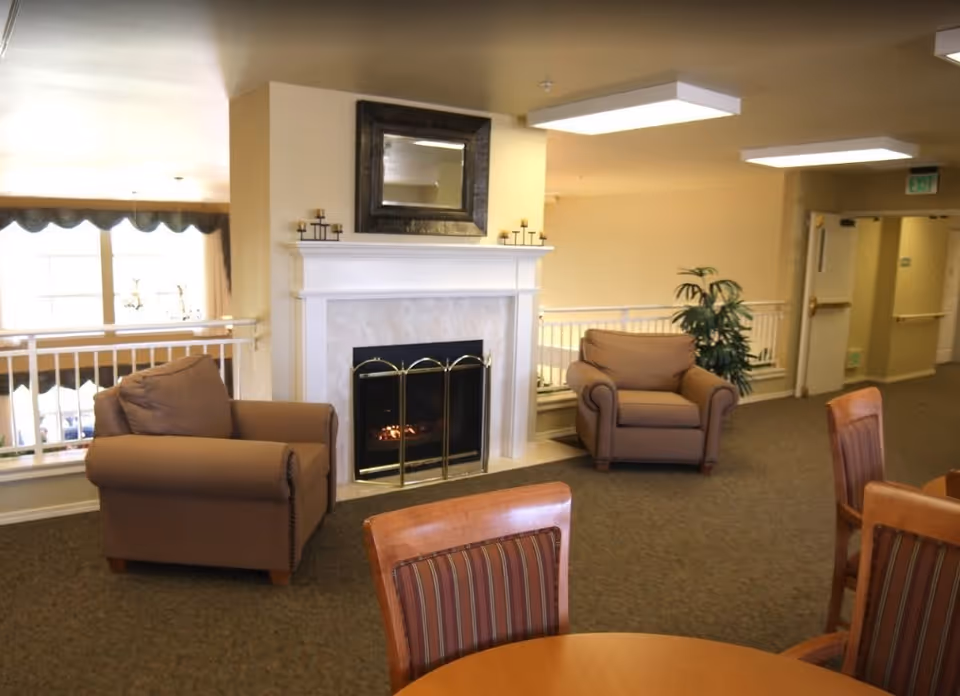A cozy common area with two brown armchairs facing a white fireplace with a black and gold screen. Above the fireplace is a large framed mirror and candle holders on the mantel. In the foreground, there are wooden chairs with striped upholstery around a round table. The room has beige walls, carpeted floor, and ceiling lights. A potted plant is placed near the armchairs, and a hallway with doors is visible in the background.