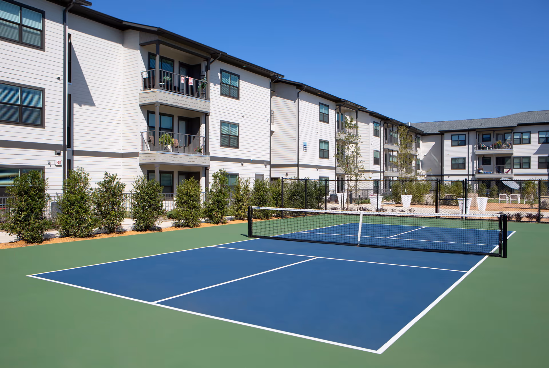 Outdoor tennis or pickleball court with a net in front of a three-story residential building with balconies and windows, surrounded by small trees and shrubs under a clear blue sky.