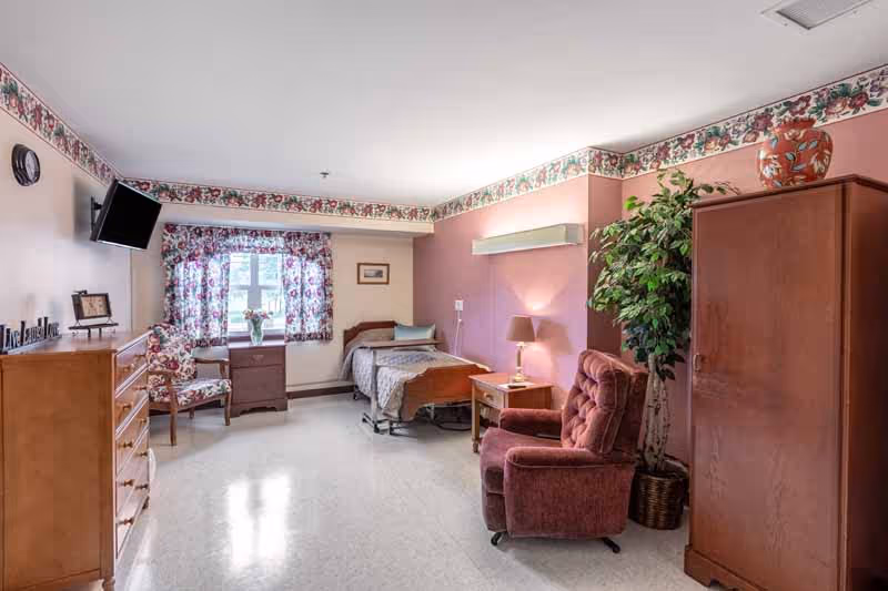 Furnished senior care bedroom with a single bed, recliner, dresser, TV, and floral curtains.