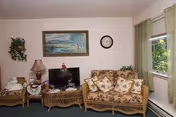 A cozy living room with wicker furniture including a sofa and two chairs with floral cushions. A small TV sits on a wicker stand, next to a lamp and decorative items. A framed landscape painting and a clock hang on the wall. There is a window with green curtains letting in natural light.