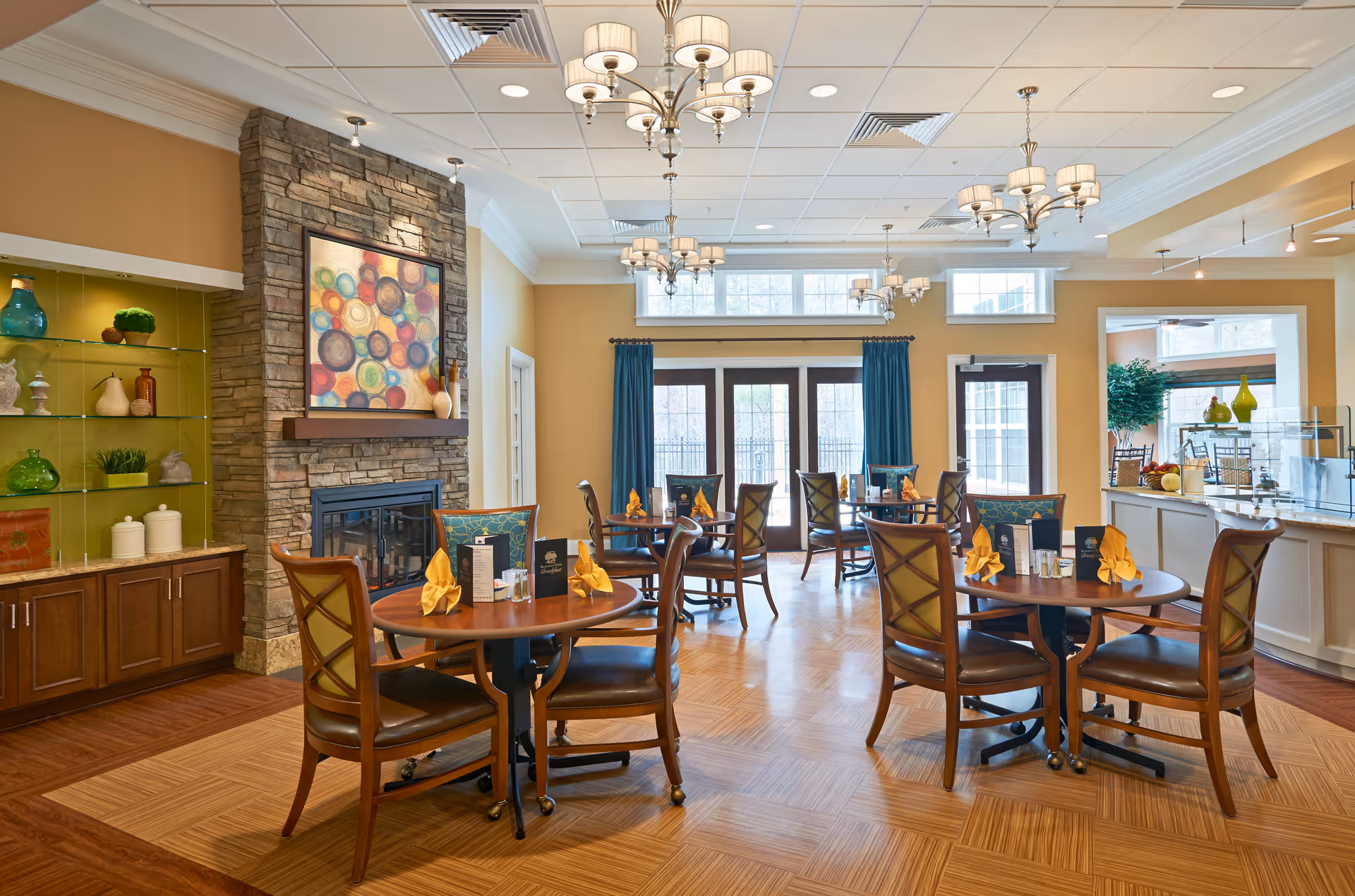 A bright and elegant dining room with several round wooden tables set with yellow folded napkins, menus, and salt and pepper shakers. The room features a stone fireplace with colorful abstract artwork above it, wooden cabinetry with decorative items, large windows with blue curtains, and multiple chandeliers hanging from the ceiling.