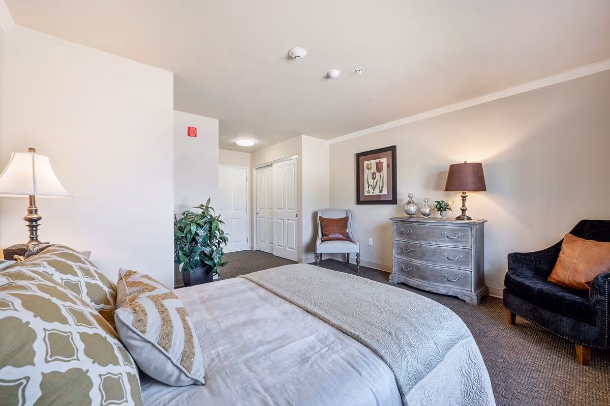 A well-lit bedroom in an assisted living facility featuring a neatly made bed with patterned pillows and a light-colored quilt. The room includes a gray dresser with decorative items and a lamp, two upholstered chairs with accent pillows, a framed floral artwork on the wall, a potted plant, and a closet with double doors. The walls are painted a soft beige color.