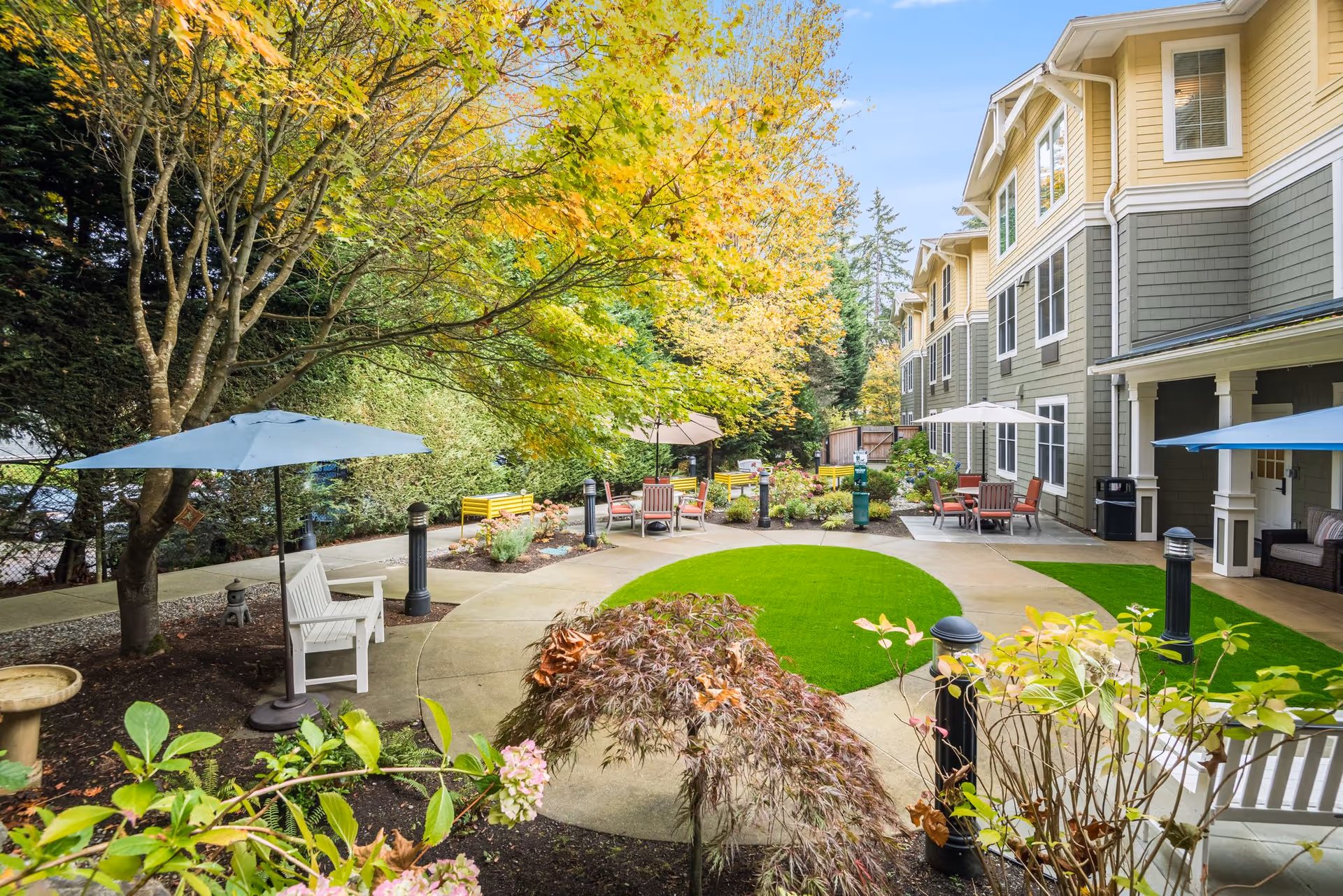 Landscaped outdoor courtyard with chairs, tables and umbrellas around a circular lawn next to a multi-story residential building.