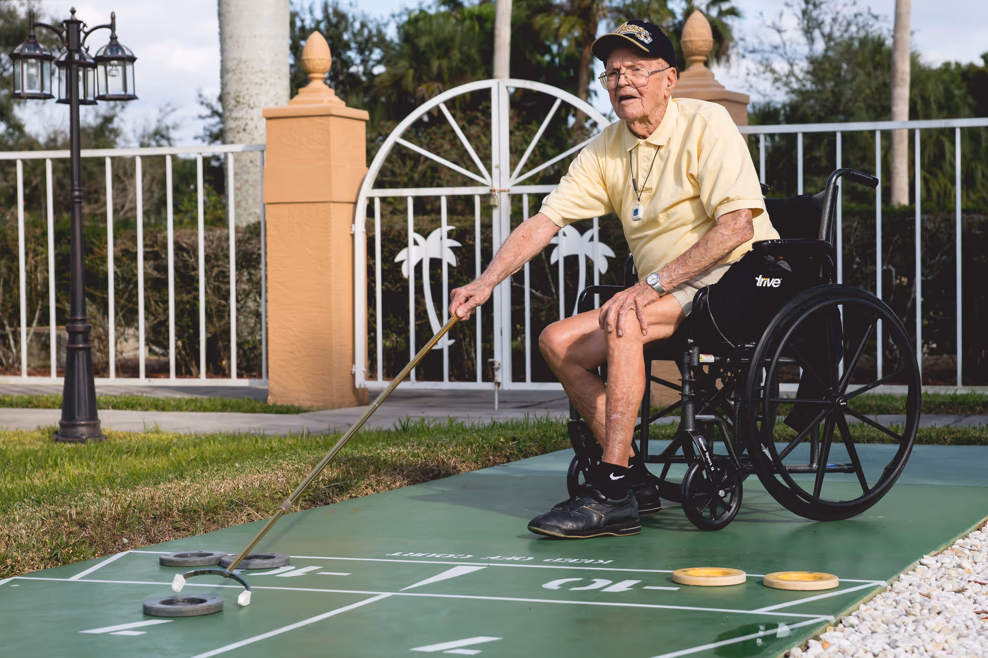 An elderly man in a wheelchair playing shuffleboard outdoors on a green shuffleboard court. He is wearing a yellow polo shirt, black shorts, black shoes, and a black cap. The background shows a white fence with palm tree designs and some greenery.