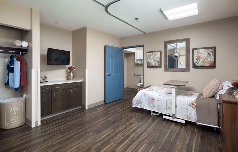 A well-lit skilled nursing facility room with a single bed covered in a floral and script patterned bedspread. The bed has a beige pillow and a brown blanket. Next to the bed is an adjustable overbed table. On the wall above the bed are two framed floral artworks and a window with a wooden frame. The room has wood-look flooring and beige walls. There is a blue door leading to a bathroom with a visible toilet and grab bars. On the opposite wall, there is a mounted flat-screen TV above a dark wood cabinet with a vase of flowers. An open closet with hanging clothes and a laundry basket labeled 'Fluff & Fold Laundromat' is visible on the left side.