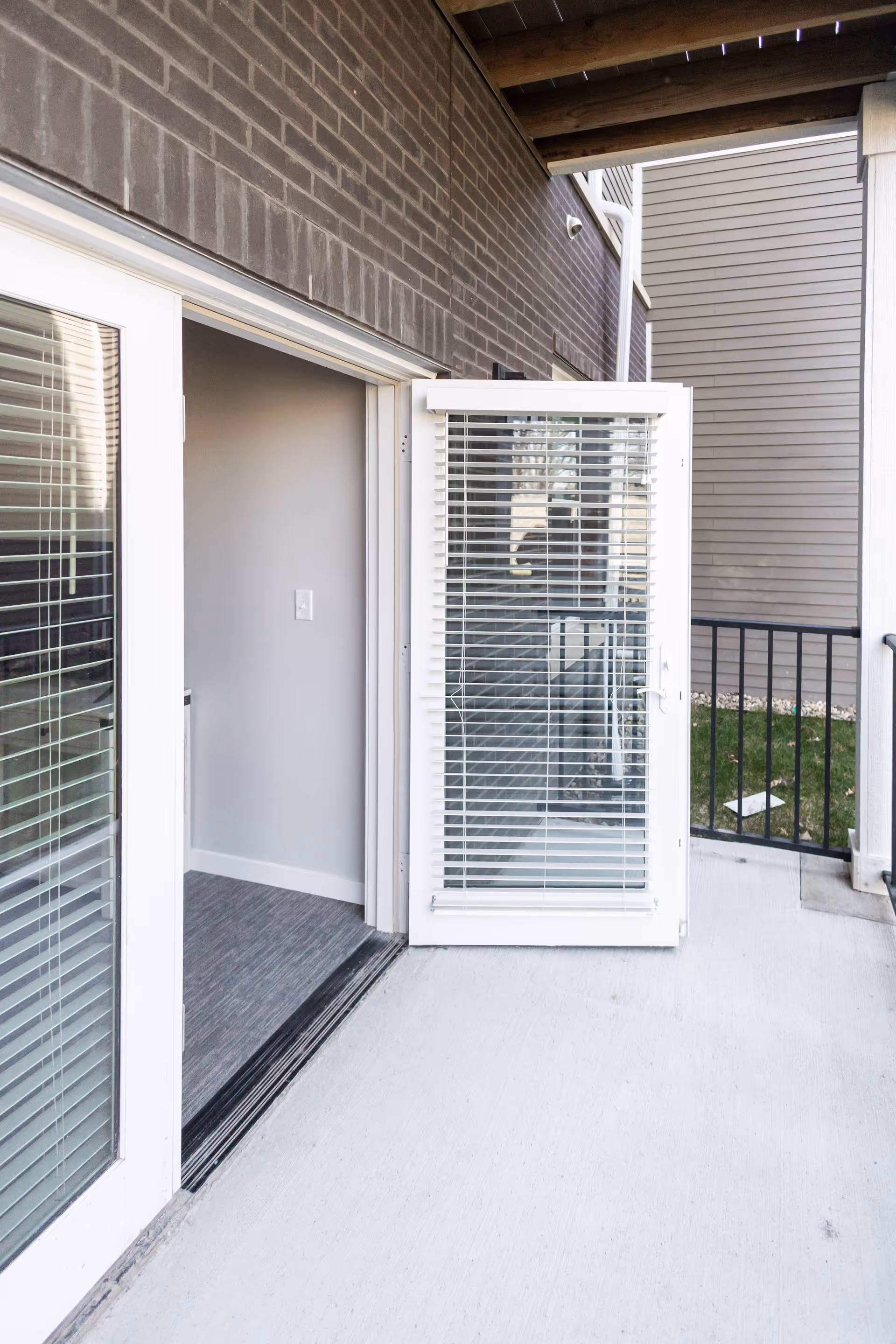 Open glass patio door with blinds leading from an interior room onto a small concrete balcony with railing and brick siding.