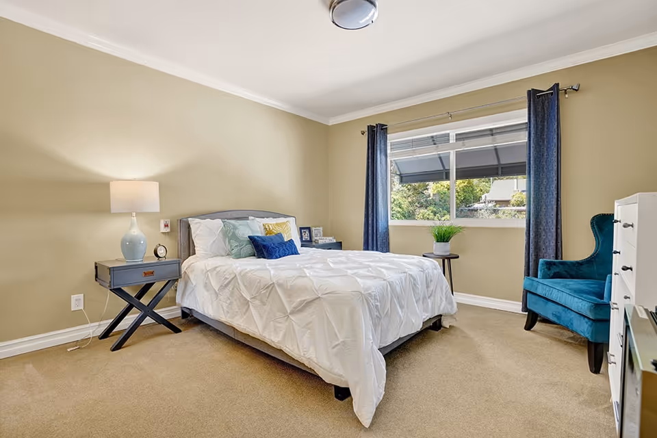 A neatly made bed with white bedding and multiple decorative pillows in a bedroom with beige walls and carpet. There is a nightstand with a lamp and clock on the left side of the bed, a window with blue curtains letting in natural light, a small round table with a plant, a blue upholstered armchair, and a white dresser on the right side.