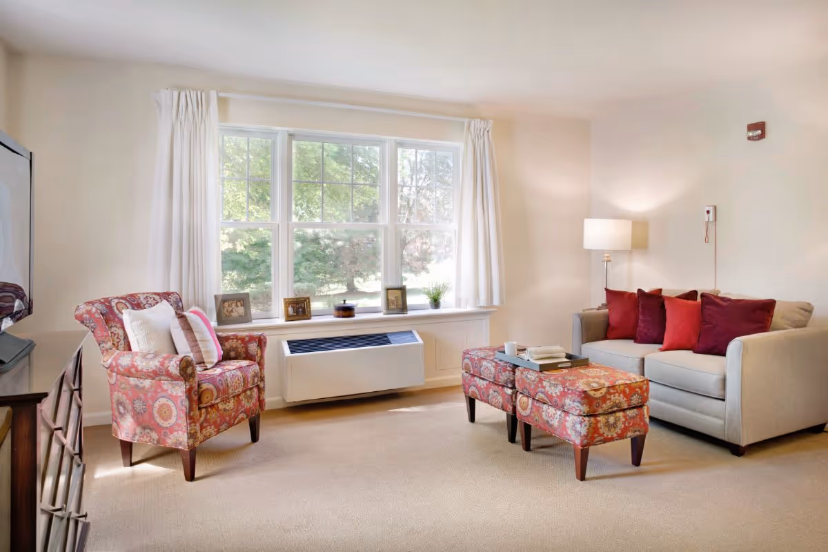 A cozy living room with a large window letting in natural light. The room features a beige sofa with red and burgundy throw pillows, a patterned armchair with cushions, and two matching patterned ottomans serving as a coffee table with books and a cup on top. A floor lamp stands next to the sofa, and framed photos and a small plant are placed on the window sill.