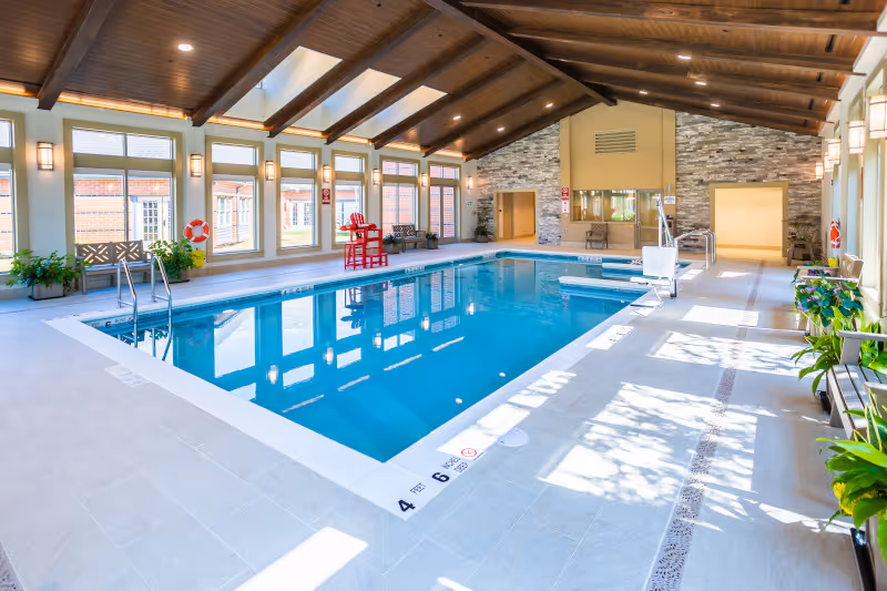 Indoor swimming pool area with clear blue water, surrounded by tiled flooring and several benches with potted plants. The room has large windows letting in natural light, a wooden beamed ceiling with recessed lighting, and a stone accent wall at the far end. There is a red lifeguard chair and life preservers on the walls.