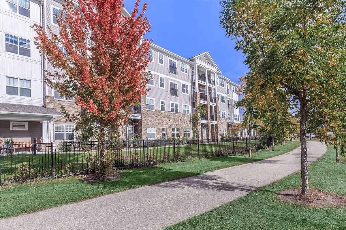 Exterior view of a multi-story assisted living facility with a stone and siding facade, surrounded by green grass and trees, including one with red autumn leaves, under a clear blue sky. A paved walking path runs alongside the building.