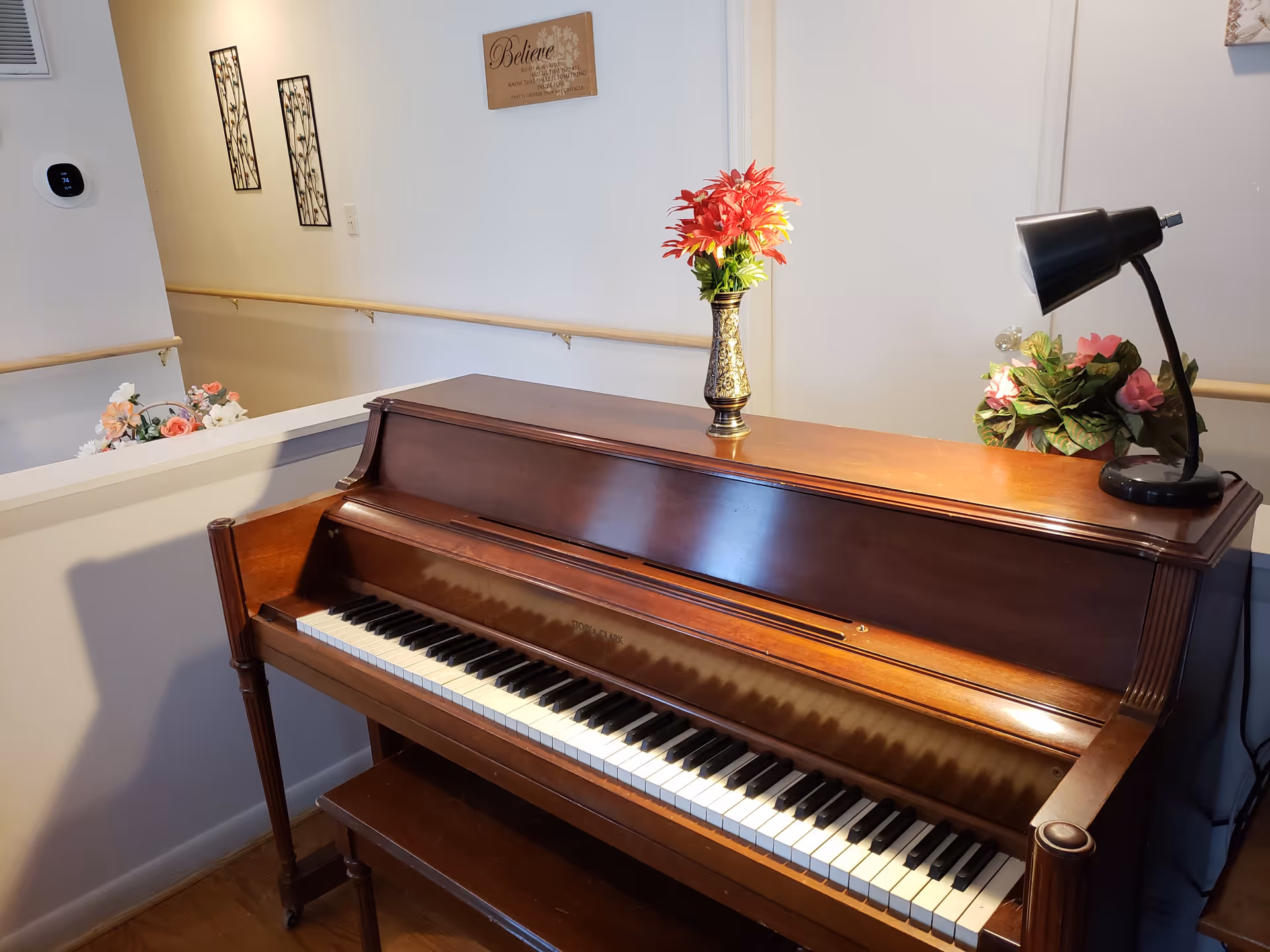 A wooden upright piano with a matching bench in a well-lit room. On top of the piano, there is a decorative vase with red flowers and a black desk lamp illuminating a small plant with pink flowers. The background shows a white wall with a wooden handrail and some wall decorations, including a framed sign that says 'Believe'.